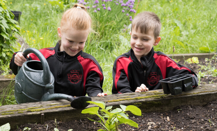 Two children in the garden, one watering plants in a bed, one holding seed pots.