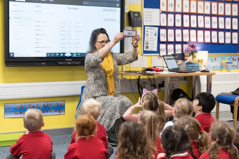 Teacher holding up the word 'Are' in front of a class sitting on the floor.